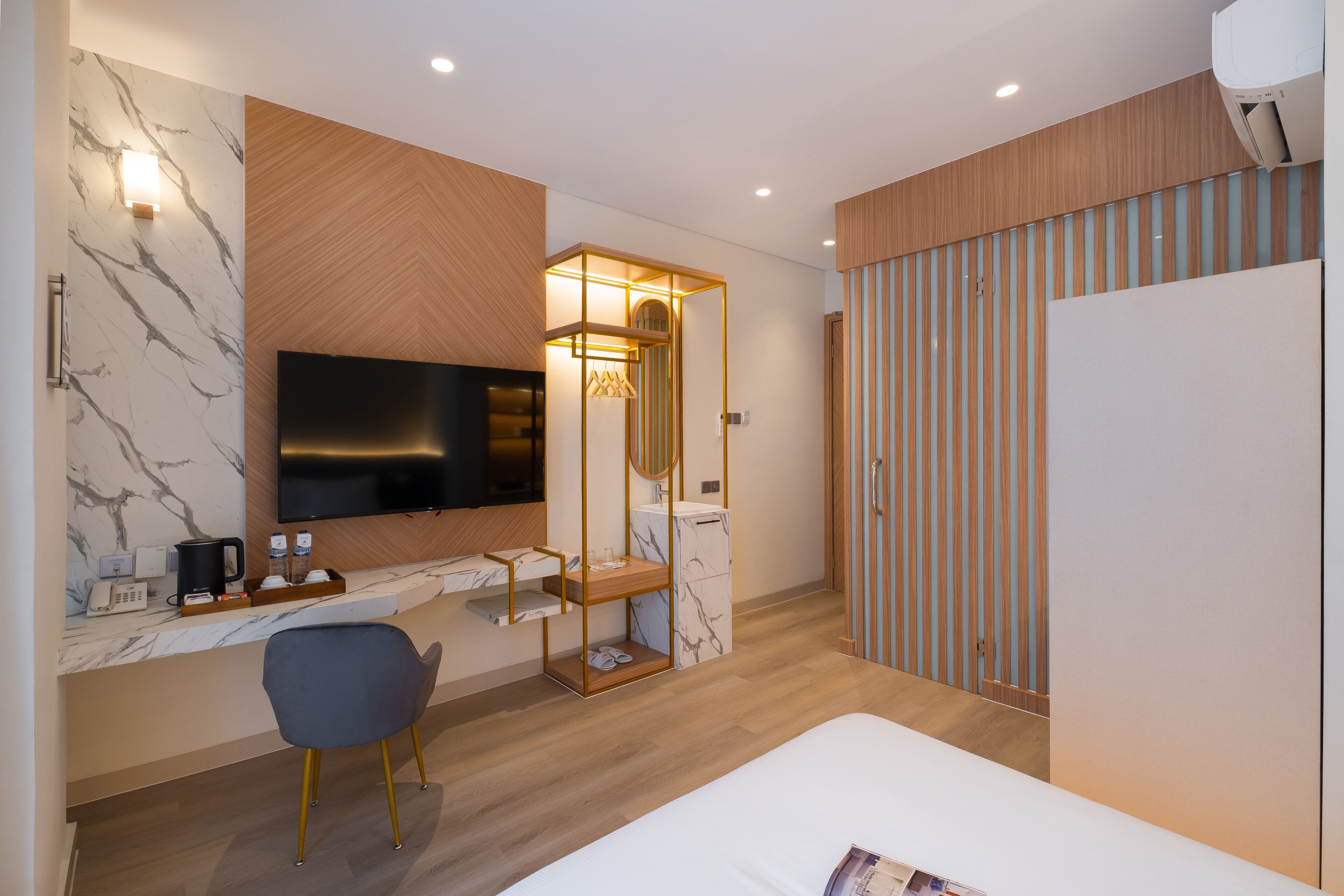 Quarto de hotel moderno com uma televisão de tela grande, uma penteadeira de madeira, parede com padrão marmorizado, cama e armário com listras verticais.