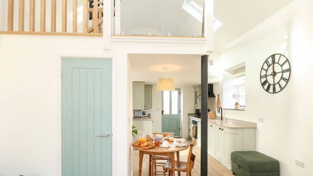 A dining area with a table and chairs, a kitchen in the background, a green door, and a clock on the wall. - Wensum Spring (Dereham)