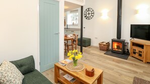 A living room with a sofa, a wooden coffee table with a vase of flowers, a fireplace, a TV, and a dining area in the background. - Wensum Spring (Dereham)