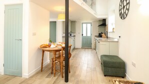 A spacious kitchen with wooden cabinets, a dining area with a table and chairs, and a green ottoman. - Wensum Spring (Dereham)