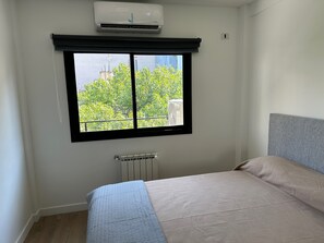 Un dormitorio con un ventanal grande, un radiador y una cama con cabecera gris.