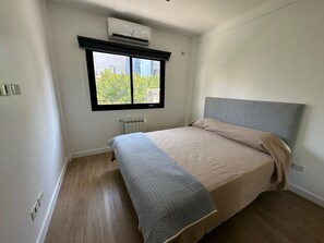 Un dormitorio con cama, radiador, equipo de aire acondicionado y ventana con vistas a la vegetación.