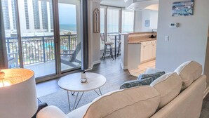 A modern living room with a sofa, a round table, and a ceiling fan. A balcony with a view of the ocean and a pool is visible through the glass doors. - Gorgeous view overlooking Currents Pool Complex and has access, 405 Brighton (Myrtle Beach)
