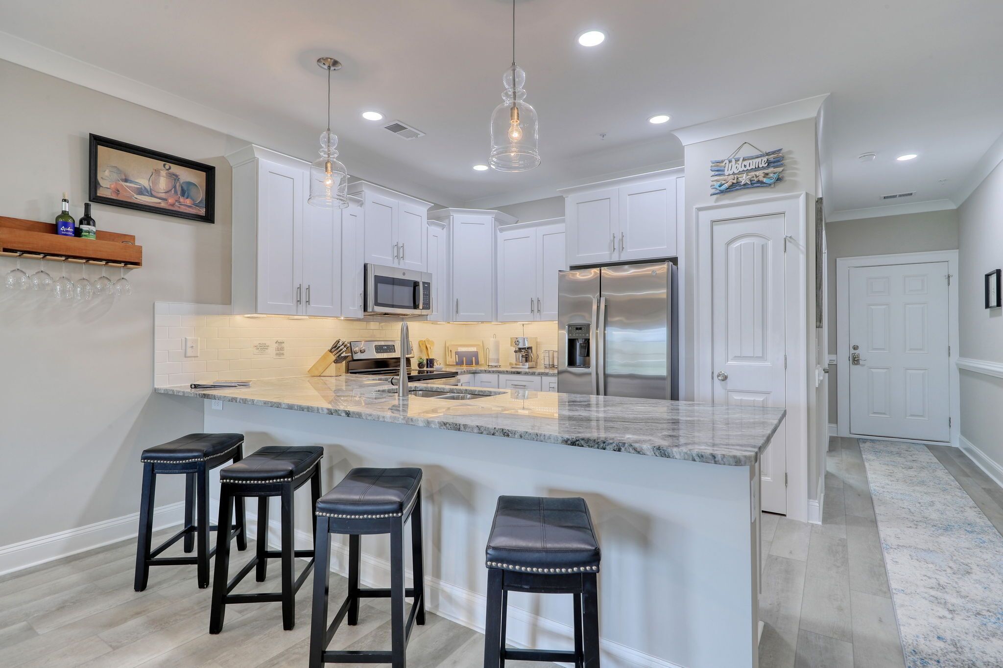 Cozinha moderna com armários brancos, uma ilha grande com banquetas, eletrodomésticos de aço inoxidável e iluminação pendente.