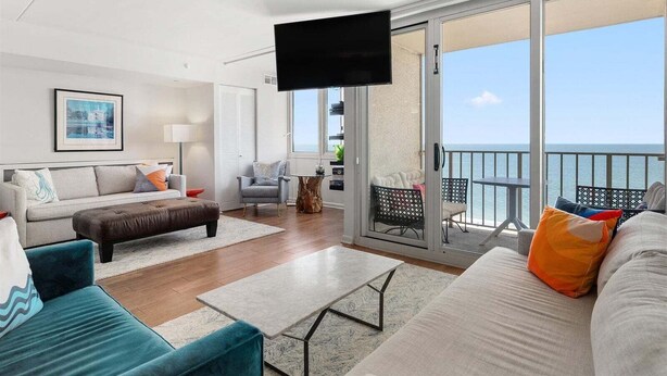 Un salon moderne comprenant un canapé, un pouf et un fauteuil, un téléviseur à écran plat, ainsi qu’un balcon offrant une vue sur la mer.