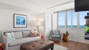 Un salon moderne avec un canapé gris, un pouf marron, un fauteuil gris et une petite table en bois. Un tableau encadré est accroché au mur, ainsi qu’une lampe sur pied et une fenêtre donnant sur la mer.