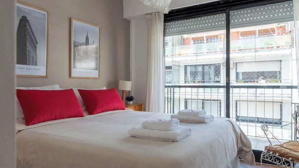 A hotel room with a large bed, red pillows, a nightstand with a lamp, and two framed pictures of buildings. - MOT Rentals + Anchorena 1400 - 2 Bd in Recoleta (Buenos Aires)