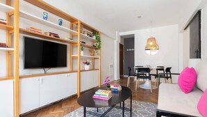 A modern living room with a flat-screen TV, wooden shelving, a dining area, and a sofa with a pink pillow. - MOT Rentals + Anchorena 1400 - 2 Bd in Recoleta (Buenos Aires)