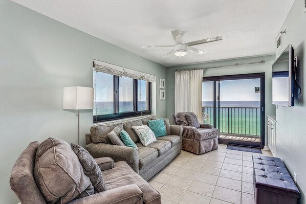 A living room with a sectional sofa, a ceiling fan, and a view of the ocean through large windows.