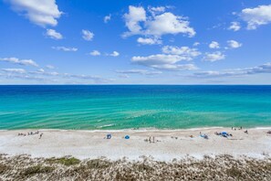 A beach with clear turquoise water and white sand, featuring several umbrellas and people relaxing.