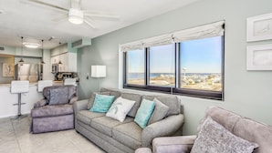 Ein Wohnzimmer mit grauem Sofa, einem kleinen Hocker und Blick auf den Strand durch große Fenster.