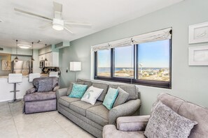 A living room with a grey sofa, a small ottoman, and a view of the beach through large windows.