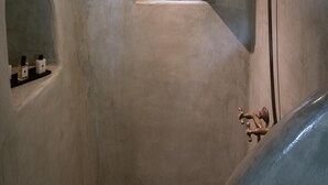 A bathroom with a small arched window, a showerhead, and a sink with a faucet. - Madame Kalitsi-ioli Standard Room (Santorini)