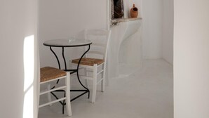 Una habitación de paredes blancas con una mesa redonda y una silla, una lámpara colgante y un pequeño ventanal con una maceta rústica.