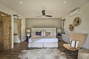Un dormitorio con una cama grande, un ventilador de techo, una puerta de madera, una silla tejida y una alfombra.