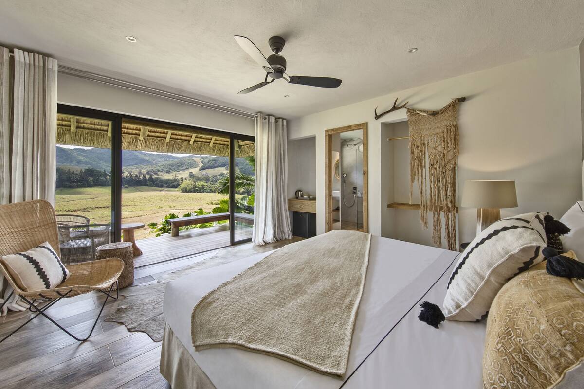 Ein Schlafzimmer mit einem großen Bett, einem Deckenventilator, Blick ins Freie, Holzboden und einem Fenster mit Vorhängen.
