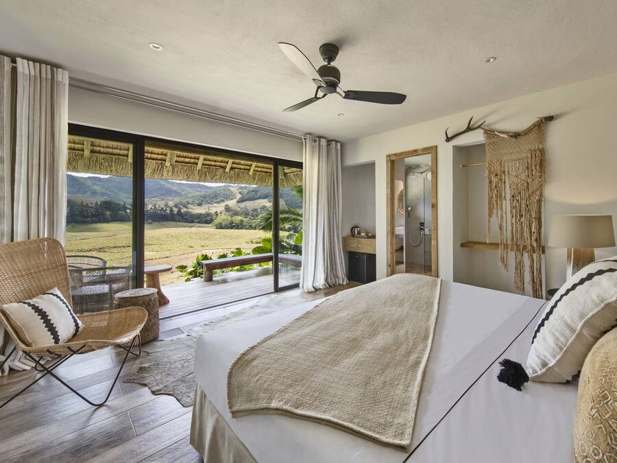 Une chambre avec un grand lit, un ventilateur de plafond, une vue sur l’extérieur, un plancher en bois et une fenêtre avec des rideaux.