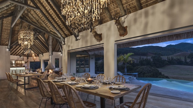 A dining area with a long wooden table set for a meal, surrounded by wicker chairs, with a view of a pool and mountains in the background. - Ferney Nature Lodge (Vieux Grand Port)
