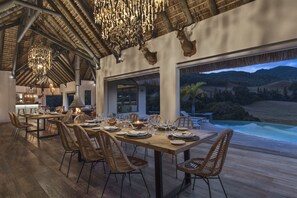 Comedor con una larga mesa de madera dispuesta para una comida, rodeado de sillas de mimbre, con vista a una piscina y montañas al fondo.