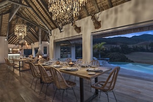Comedor con una larga mesa de madera dispuesta para una comida, rodeado de sillas de mimbre, con vista a una piscina y montañas al fondo.