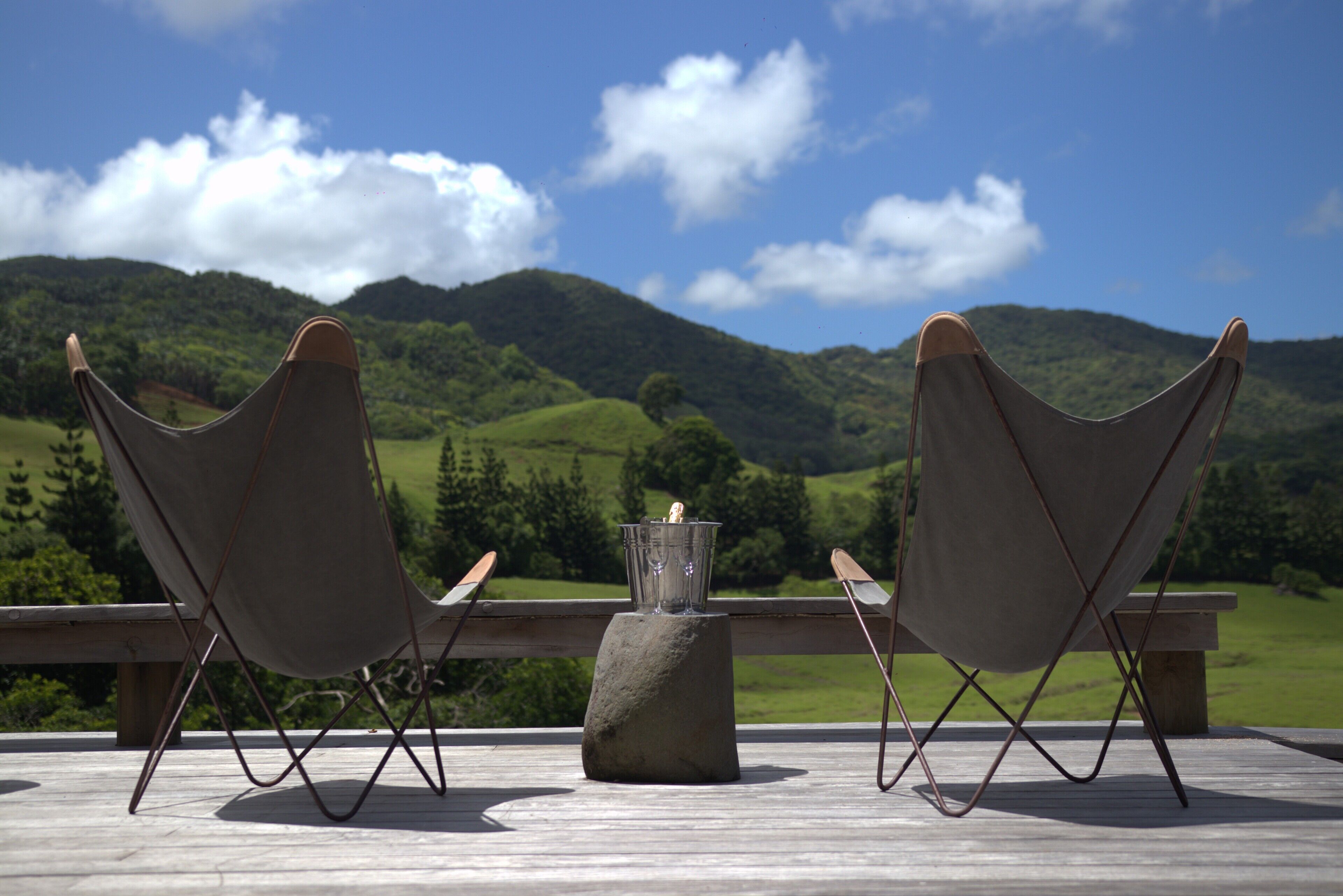 Dos sillas sobre una terraza de madera con una mesa de piedra en el centro, con vistas a un paisaje verde y colinas.