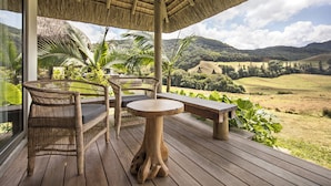 A wooden deck with wicker furniture and a table, overlooking a lush landscape with hills and a clear sky. - Ferney Nature Lodge (Vieux Grand Port)