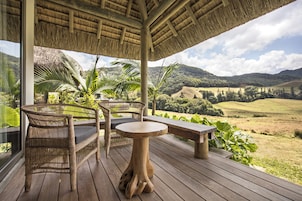 Terraza de madera con mobiliario de mimbre y una mesa, con vistas a un paisaje exuberante con colinas y cielo despejado.