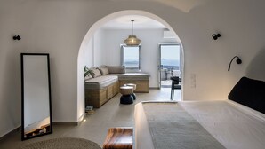 A modern bedroom with a large bed, a seating area with a sofa and cushions, a dining table, and a view of the sea through an open window. - WeSense by Caldera Houses (Santorini)