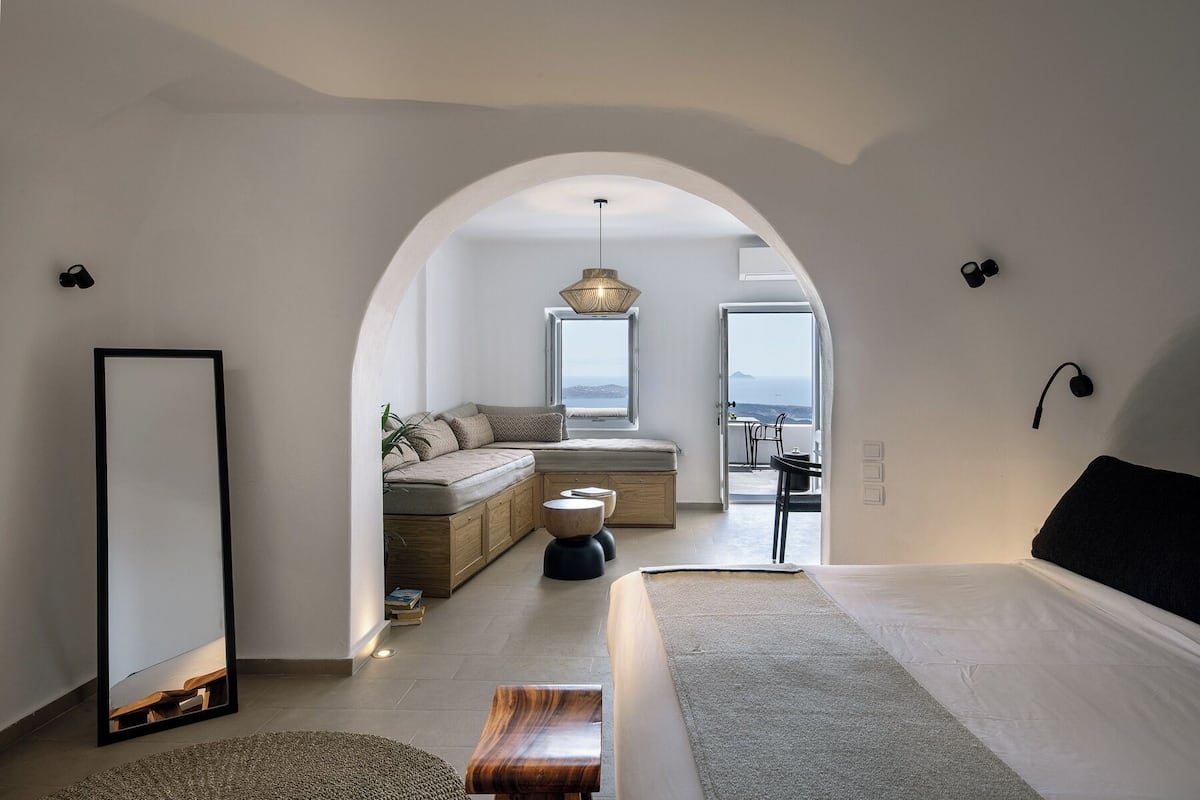 A modern bedroom with a large bed, a seating area with a sofa and cushions, a dining table, and a view of the sea through an open window.