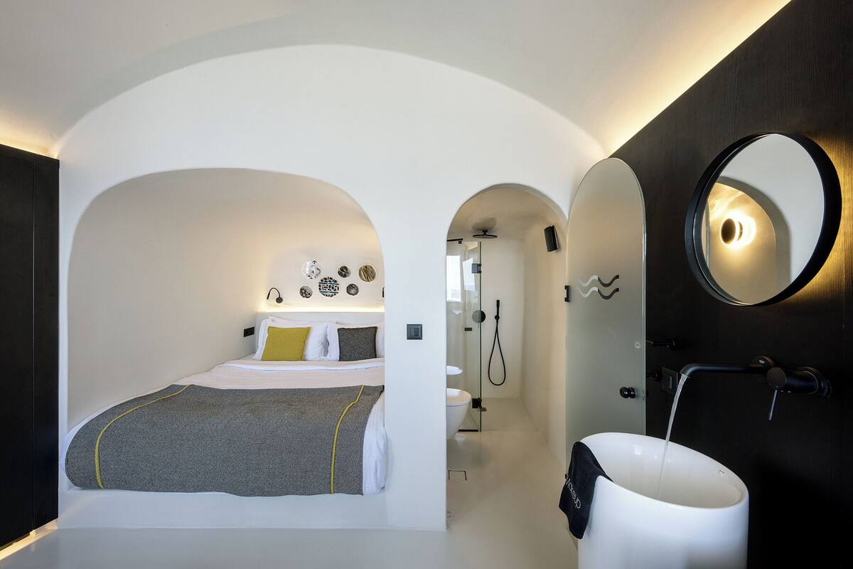 A modern bedroom with a bed, a bathroom with a sink and mirror, and an arched doorway leading to a shower area.