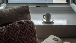 A cozy reading nook with a view of a coastal landscape, a small table with a cup, and a book on a windowsill. - WeSense by Caldera Houses (Santorini)