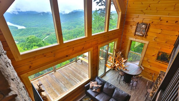 Une cabane en bois aux plafonds hauts, dotée d’une grande fenêtre donnant sur une vallée boisée. À l’intérieur, on trouve un canapé en cuir, un pouf rond et une table à manger avec des chaises.