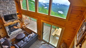 Un intérieur de chalet chaleureux avec une cheminée en pierre, des fauteuils en cuir et de grandes fenêtres offrant une vue sur les montagnes.