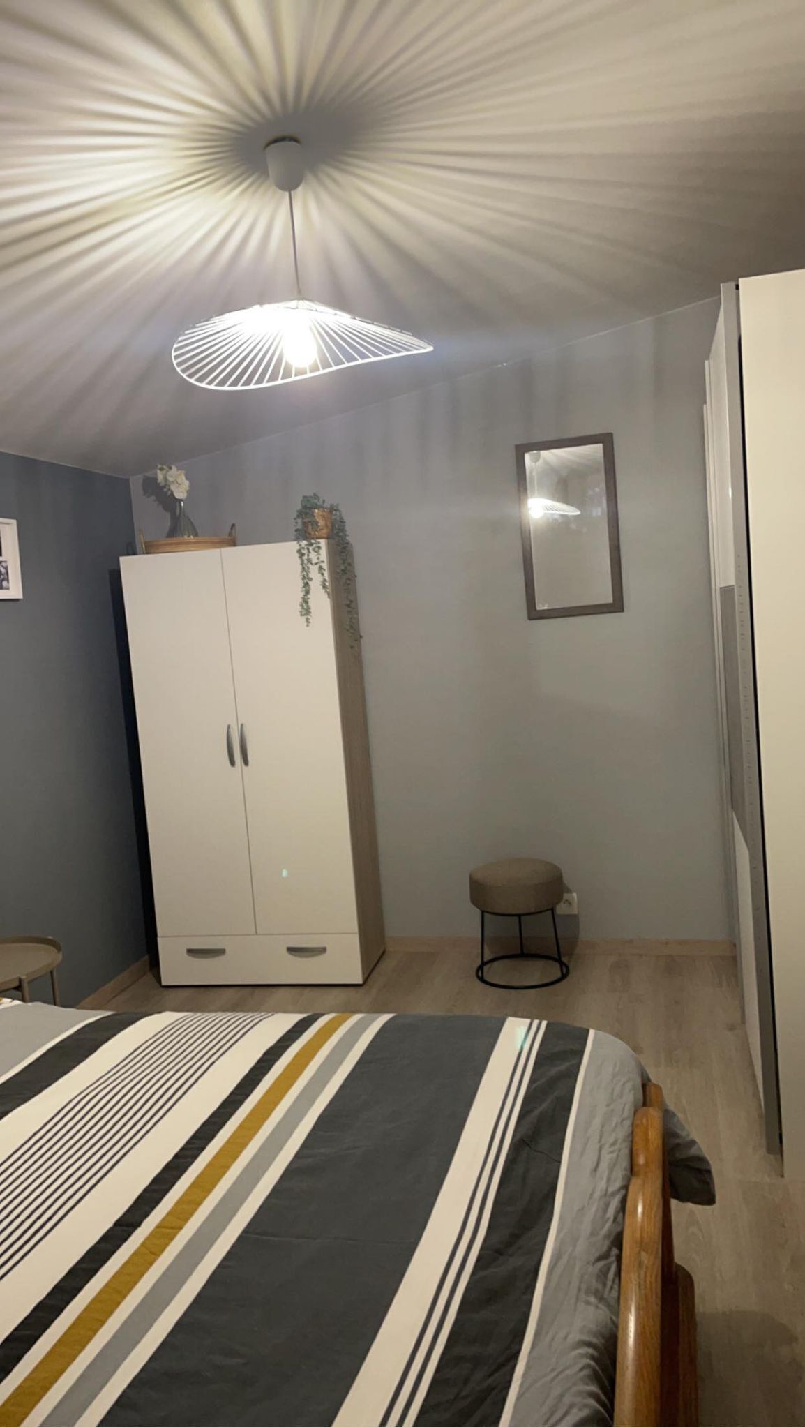 Een slaapkamer met een bed, een kledingkast, een klein krukje, een spiegel en een plafondlamp.
