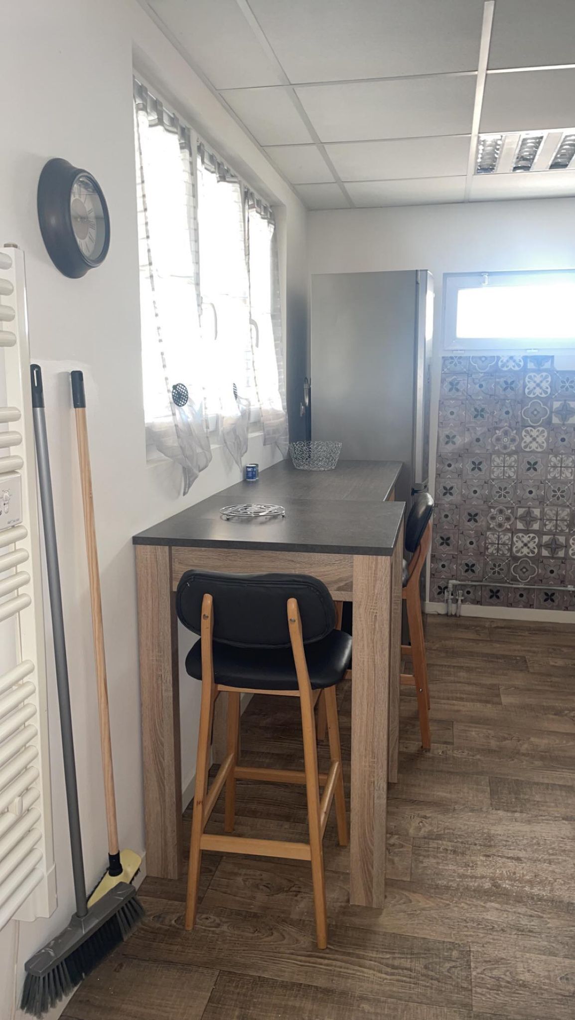 Een compacte, moderne keuken met een houten tafel en twee stoelen, een raam met dunne gordijnen en een betegelde spatwand.