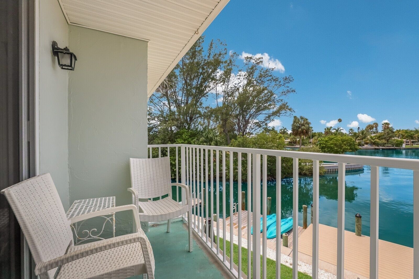 A balcony with two wicker chairs, a small table, and a view of a swimming pool with a wooden deck and palm trees.