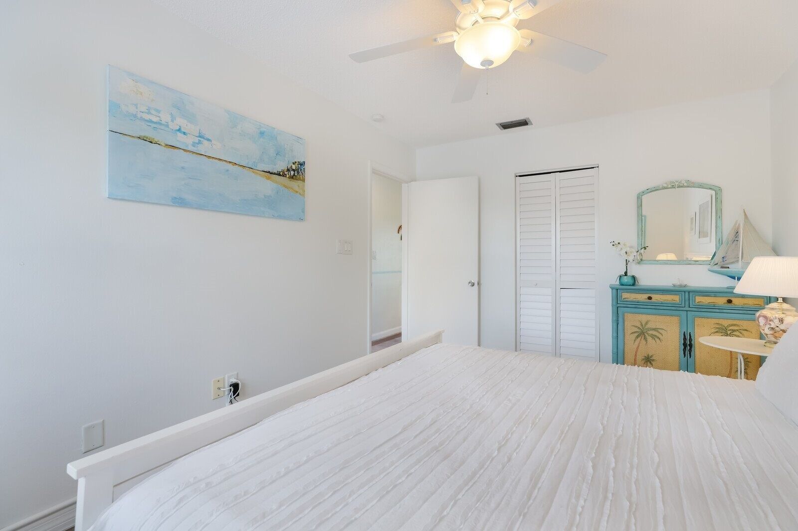 A bedroom with a bed, a ceiling fan, a painting, a wardrobe, and a vanity with a mirror.