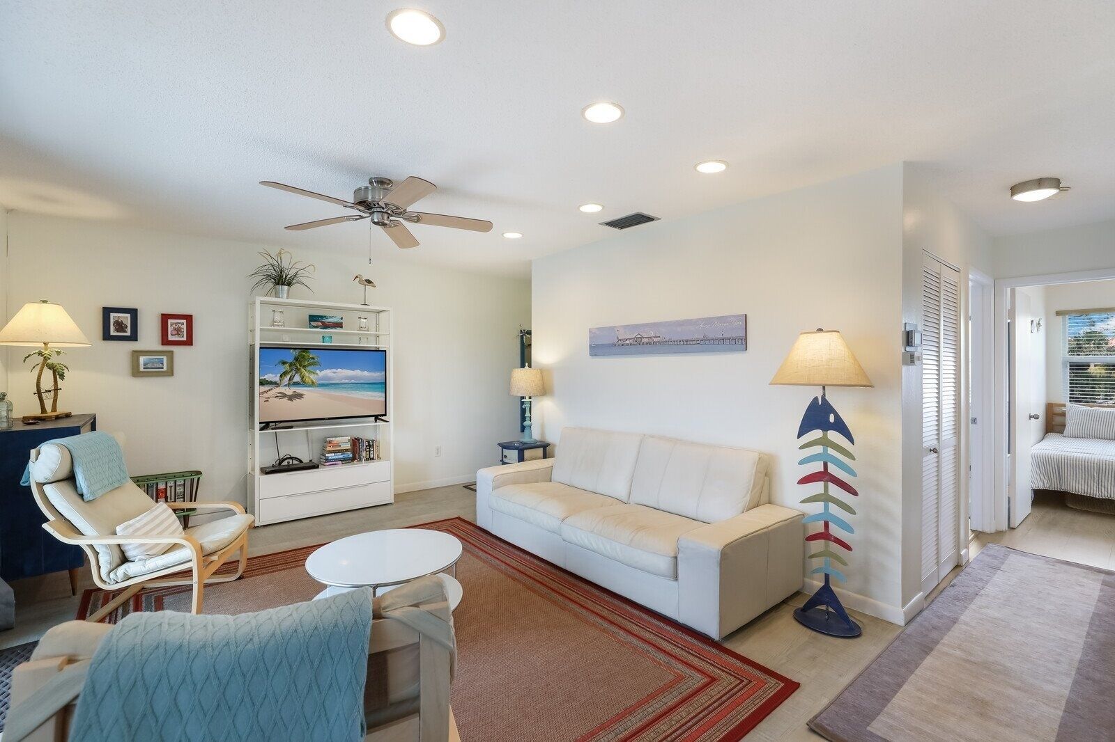 A living room with a white sofa, a white armchair, a small round table, a ceiling fan, a bookshelf with a TV, and a beach-themed painting.