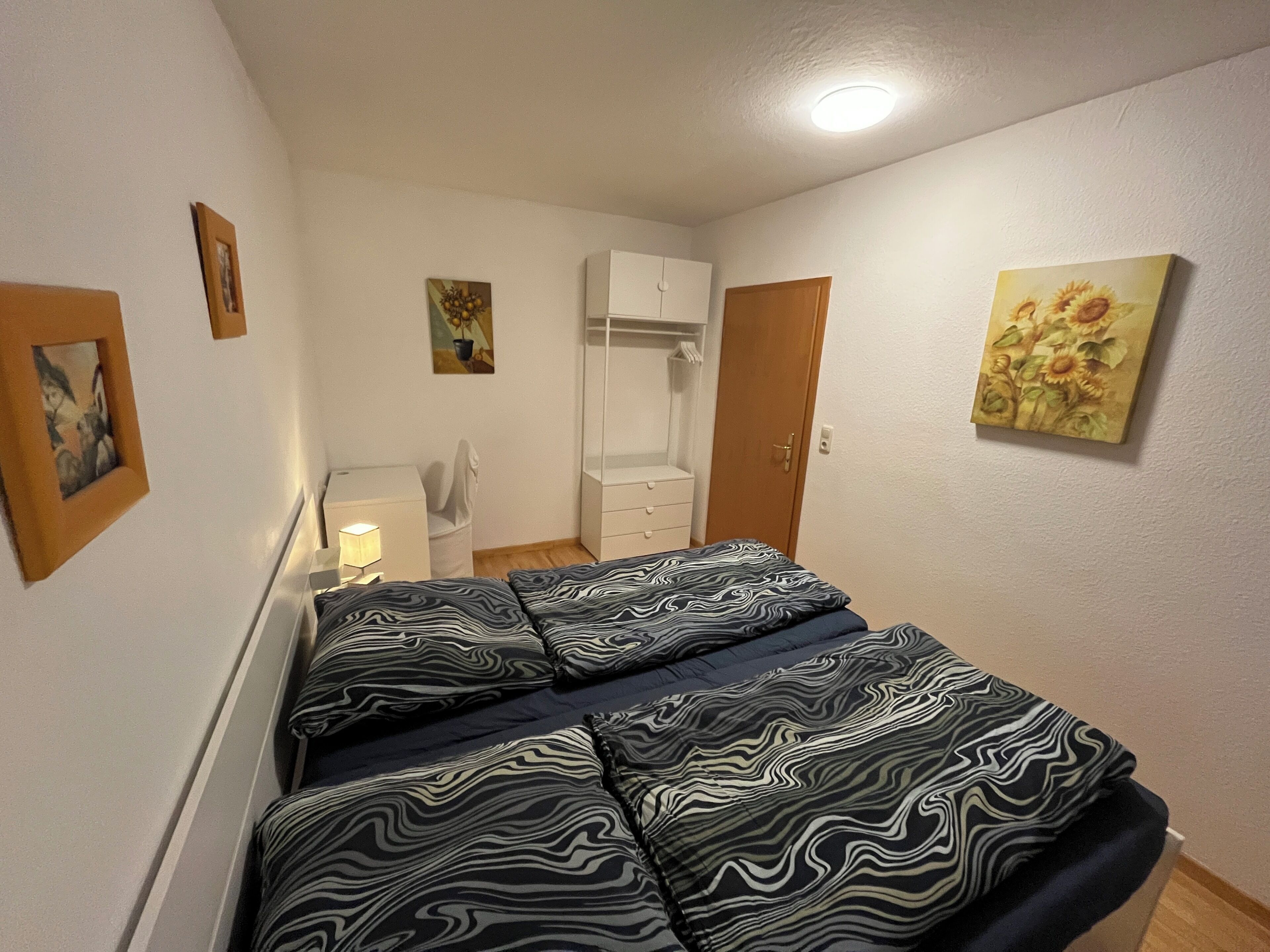 Een slaapkamer met een bed, een kledingkast, een bureau met een lamp en een schilderij aan de muur.