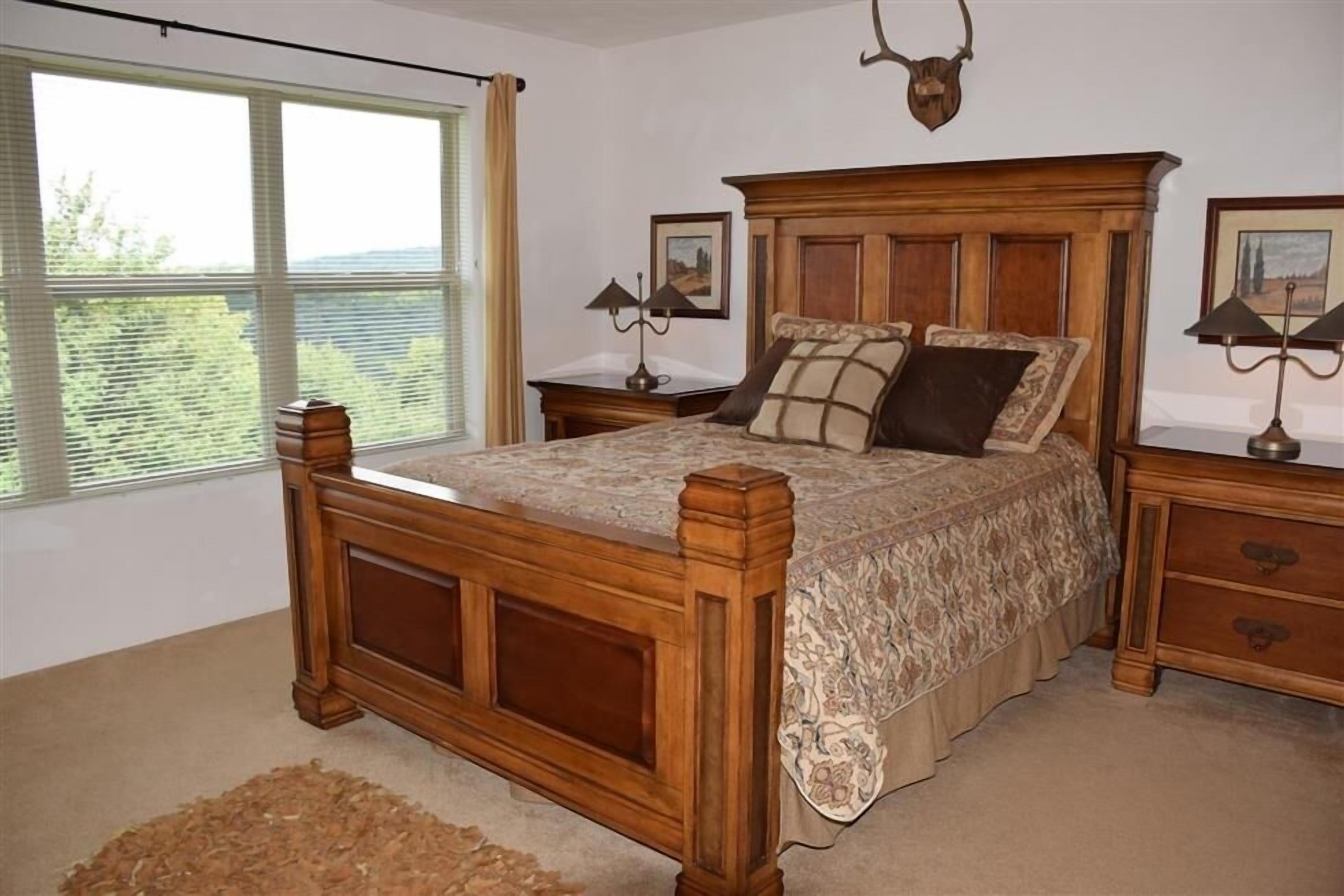 A bedroom with a wooden bed, two bedside tables, a large window with blinds, and a ceiling-mounted light fixture.