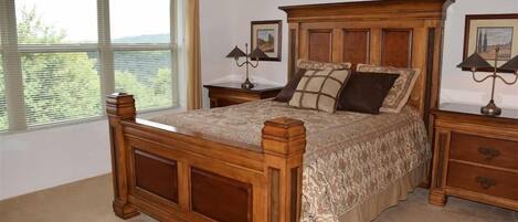 A bedroom with a wooden bed, two bedside tables, a large window with blinds, and a ceiling-mounted light fixture.