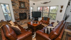 A living room with leather sofas, a stone fireplace, a flat-screen TV, and large windows with a view of trees. - Seven Springs 4 BR Premium Condo, Mountain Views! by RedAwning (Champion)