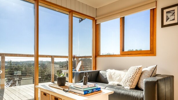 A modern living room with a sofa, a coffee table with books, and large windows offering a view of the city. - Moonah Horizon (Rye)