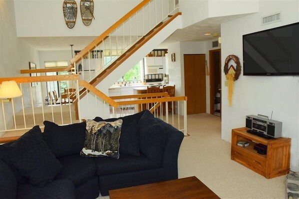 Een woonkamer met een donkergrijze bank, een houten salontafel, een televisie op een houten dressoir en een trap naar een andere kamer.