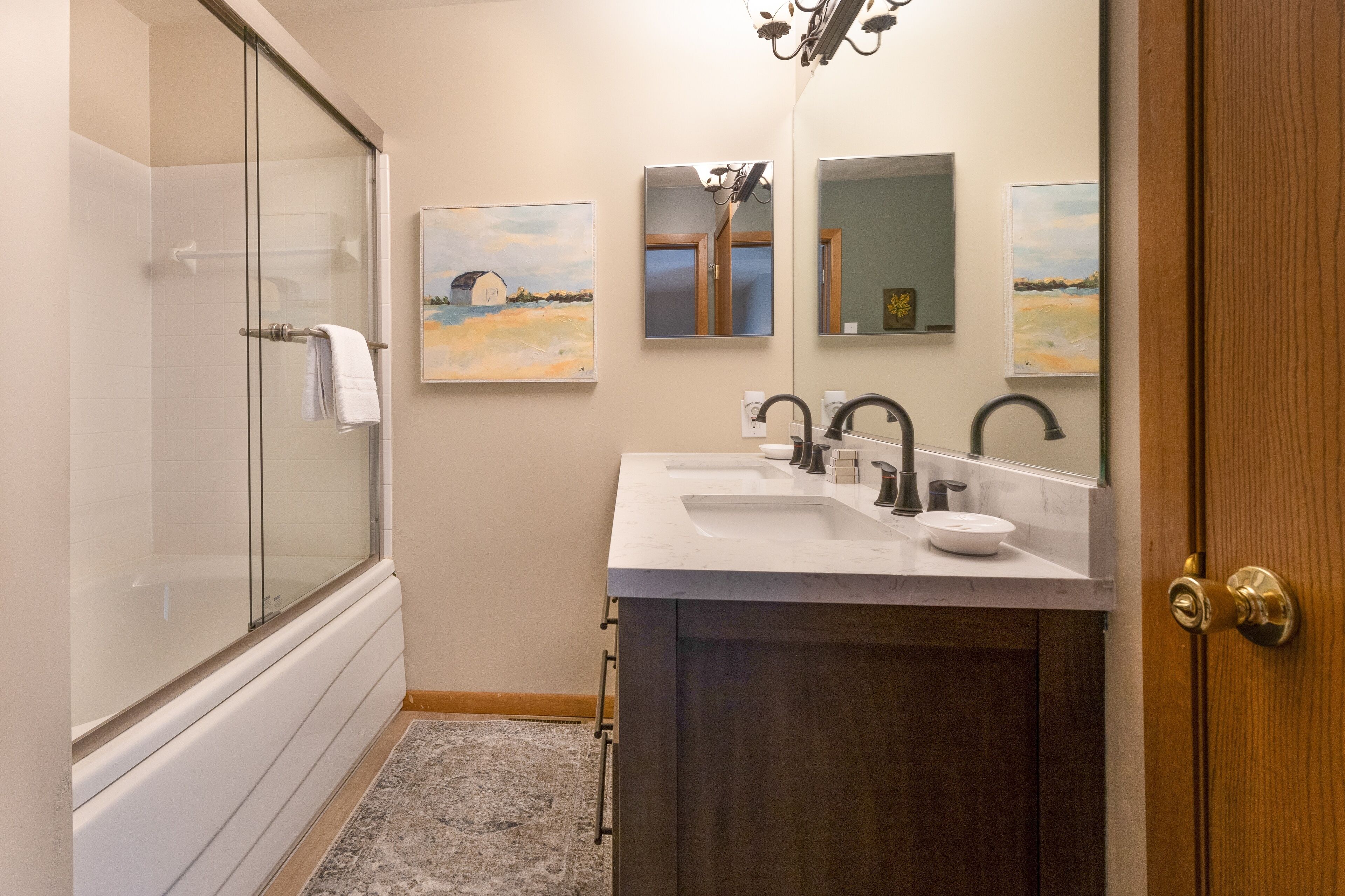 Une salle de bain moderne dotée d’une douche à parois de verre, d’un meuble-lavabo à deux vasques et de trois œuvres d’art encadrées accrochées au mur.