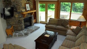 A cozy living room with a stone fireplace, plaid sofas, a coffee table, and a TV mounted on the wall.