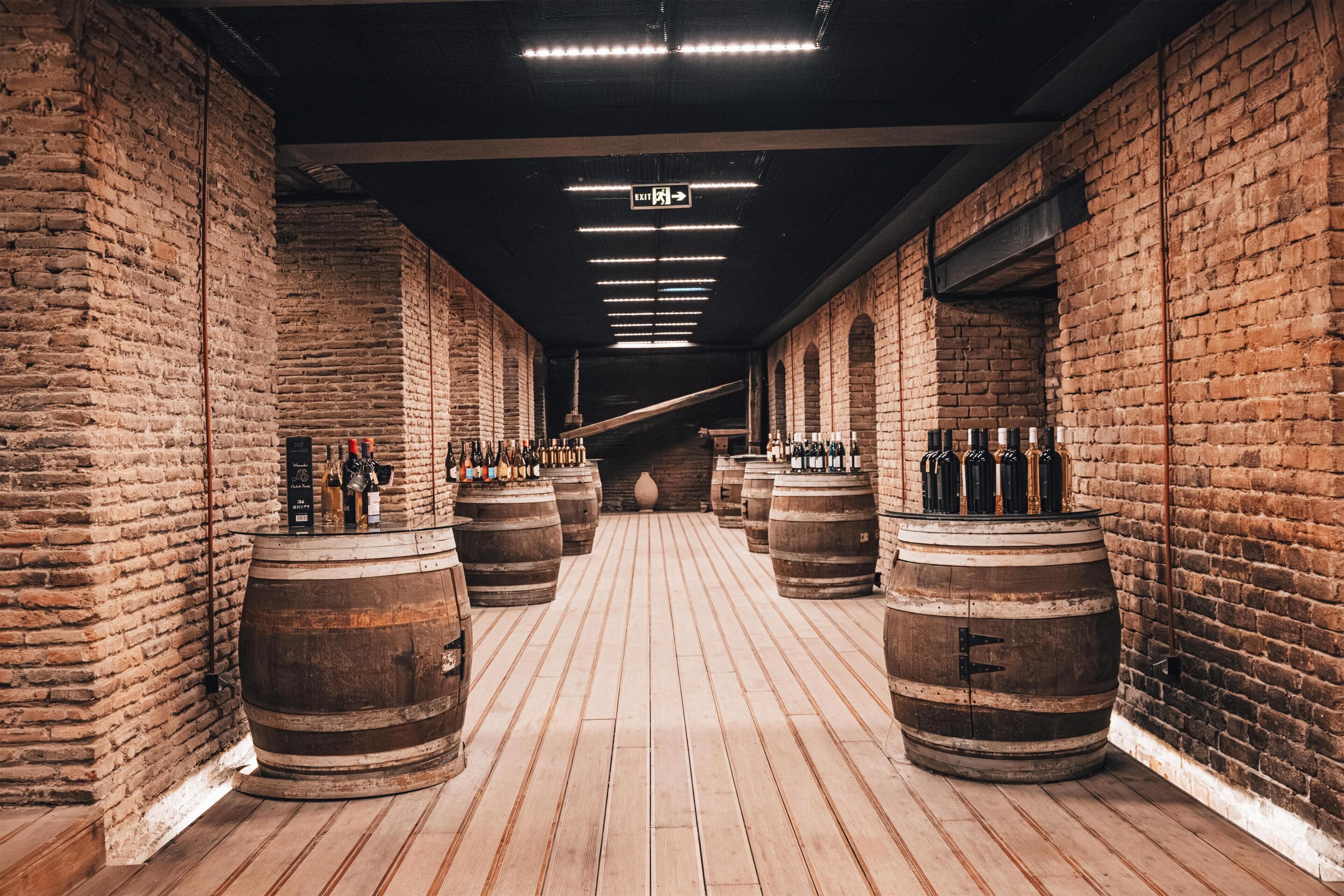 Un couloir aux murs de briques, agrémenté de barils et de bouteilles en bois, évoquant une cave à vin ou une salle de dégustation.