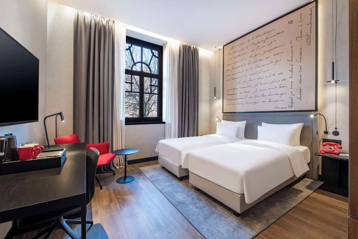 A hotel room with two beds, a desk, a chair, a TV, and a large window with curtains.