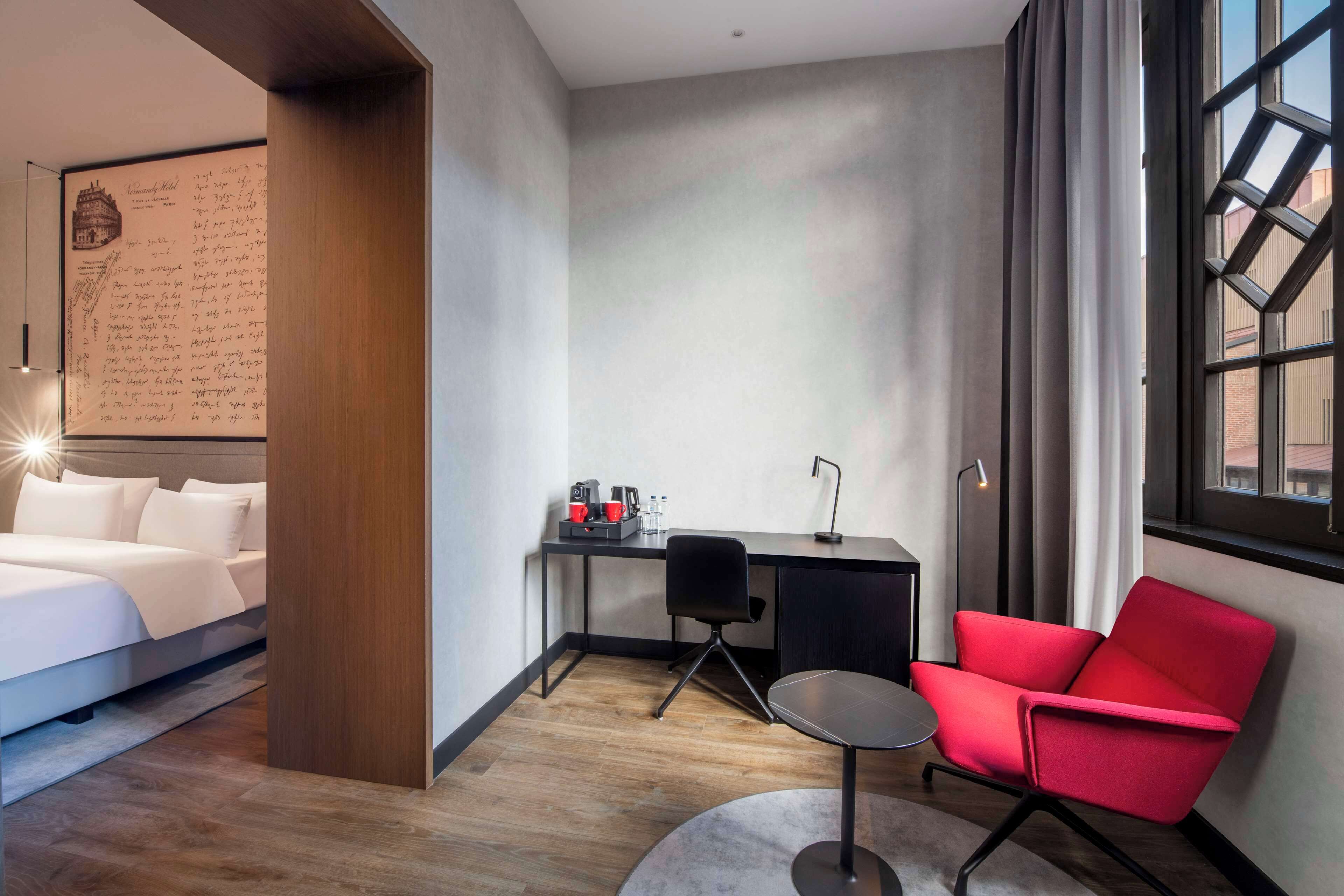 A hotel room with a bed, a desk with a chair, a red chair, a map on the wall, and a window with curtains.