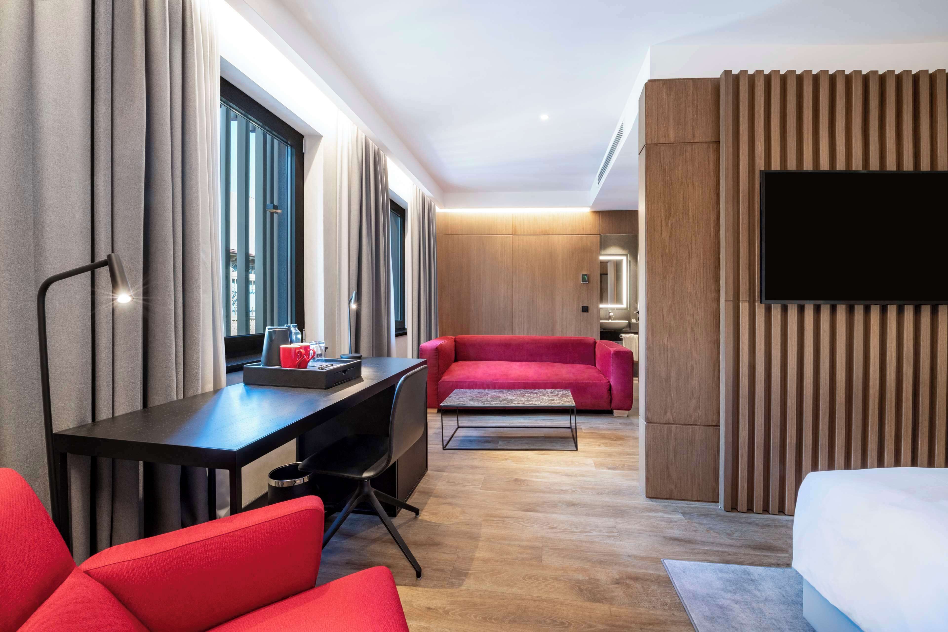 A modern hotel room with a desk, red sofa, and a TV mounted on the wall.